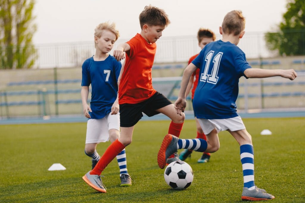 Young Football Players Kicking Ball on Soccer Field. Soccer Horizontal Background. Youth Junior Athletes in Red and Blue Soccer Shirts